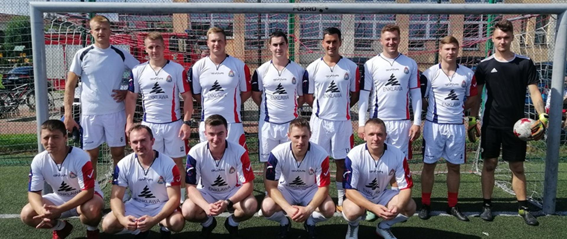 Zdjęcie przedstawia 13 funkcjonariuszy KP PSP Skarżysko-Kamienna pozujących do zdjęcia po turnieju Tevor Cup. Zawodnicy stoją w bramce, a bramkarz po prawej stronie trzyma w ręku piłkę. W tle widać siatki zabezpieczające oraz za nimi budynek szkoły podstawowej. Tuż przed budynkiem stoi samochód strażacki