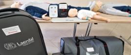 Zdjęcie przedstawiają zestaw do ćwiczeń i nauki kwalifikowanej pierwszej pomocy. Na zdjęciu widać trzy manekiny naturalnej wielkości dorosłych, dzieci oraz mniejszy manekin niemowlęcia, leżące na stole, używane do ćwiczeń resuscytacji krążeniowo-oddechowej (RKO). Obok manekinów znajdują się urządzenia treningowe AED (automatyczny defibrylator zewnętrzny). Na pierwszym planie widoczne są również torby transportowe z logo Laerdal, firmy produkującej sprzęt medyczny i szkoleniowy. torbach i urządzeniach widoczne są etykiety z napisem „ Zakup dofinansowano ze środków Samorządu Województwa Mazowieckiego w ramach zadania Wsparcie Komend Powiatowych i Miejskich PSP”. Tło stanowi jasna ściana i fragment wnętrza pomieszczenia świetlicy strażackiej PSP.