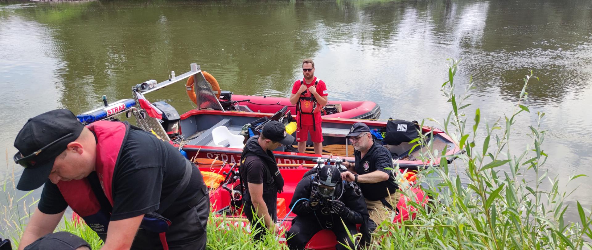 na zdjęciu widać strażaków, którzy pomagają w przygotowaniu strażaka płetwonurka do rozpoczęcia akcji. Widać dwie łódki straży pożarnej oraz łódkę WOPR, na której stoi ratownik