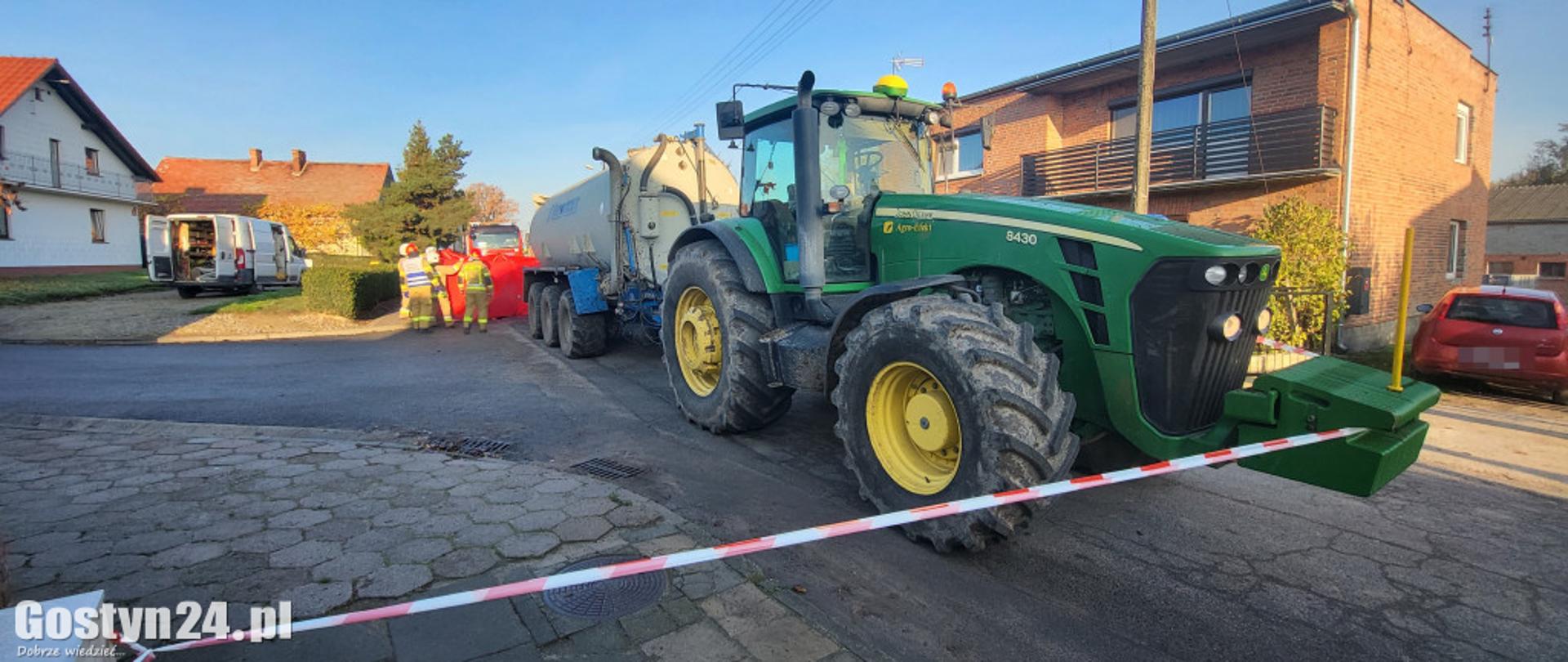 Zdjęcie przedstawia ciągnik rolniczy z przyczepą. W tle stoją służby ratownicze.