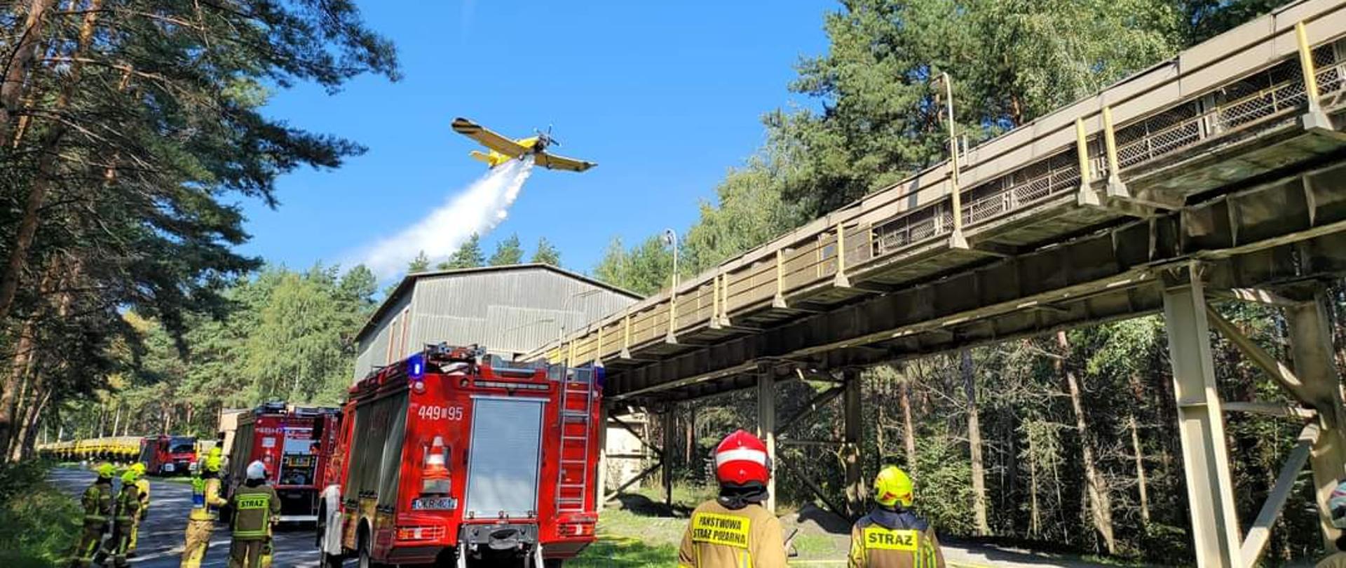 Przy budynku do którego prowadzi taśmociąg biegnący wzdłuż ściany lasy , na drodze ustawione 2 pojazdy pożarnicze. Wokół stojący strażacy obserwujący przelatujący nad lasem i budynkiem samolot gaśniczy "dromader" - dokonujący zrzutu wody. Strażacy w ubraniach specjalnych. 