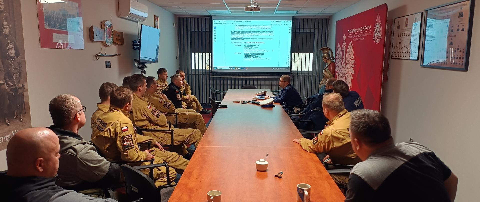 Zdjęcie przedstawia spotkanie służbowe odbywające się w sali konferencyjnej Państwowej Straży Pożarnej. Liczna grupa funkcjonariuszy w piaskowych i ciemnych mundurach siedzi wzdłuż długiego, drewnianego stołu, skupiając uwagę na prezentacji wyświetlanej z projektora. Na ekranie widoczny jest dokument tekstowy, prawdopodobnie dotyczący przepisów lub procedur, co wskazuje na szkolenie lub odprawę operacyjną.
Wystrój pomieszczenia podkreśla tożsamość formacji – na czerwonej ściance widnieje duże godło Polski oraz nazwa jednostki, a w rogu sali stoi figura św. Floriana. Na ścianach zawieszono historyczne fotografie oraz tablice informacyjne, a na stole, obok kubków z napojami i telefonów, leżą czapki mundurowe. Atmosfera spotkania wydaje się merytoryczna i profesjonalna.

