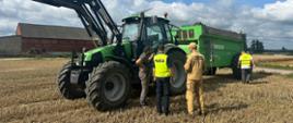 Na zdjęciu widać czerwony traktor marki Ursus stojący na polu. Obok niego znajdują się trzy osoby. Wszystkie osoby stoją na polu, jest ładna pogoda. Osoby rozmawiają ze sobą. W tle jest maszyny rolnicze i zielone pola.