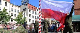 Maszt z flagą narodową w trakcie podnoszenia przez żołnierzy RP. Rzecz dzieje się na rynku, w dali tłum obserwatorów.