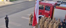 Plac przed komendą. Na tle wozu strażackiego w dwuszeregu stoją strażacy w umundurowaniu służbowym w kolorze piaskowym oraz koszarowym. Na przeciwko, przy biało-czerwonym maszcie flagowym znajduje sie poczet flagowy w umundurowaniu wyjściowym. Flagowy podnosi flagę, natomiast dowódca pocztu i asystujący salutują. Z lewej strony w pozycji zasadniczej w umundurowaniu wyjściowym stoi zastępca komendanta powiatowego, który również oddaje honor. Za nim droga i budynki.