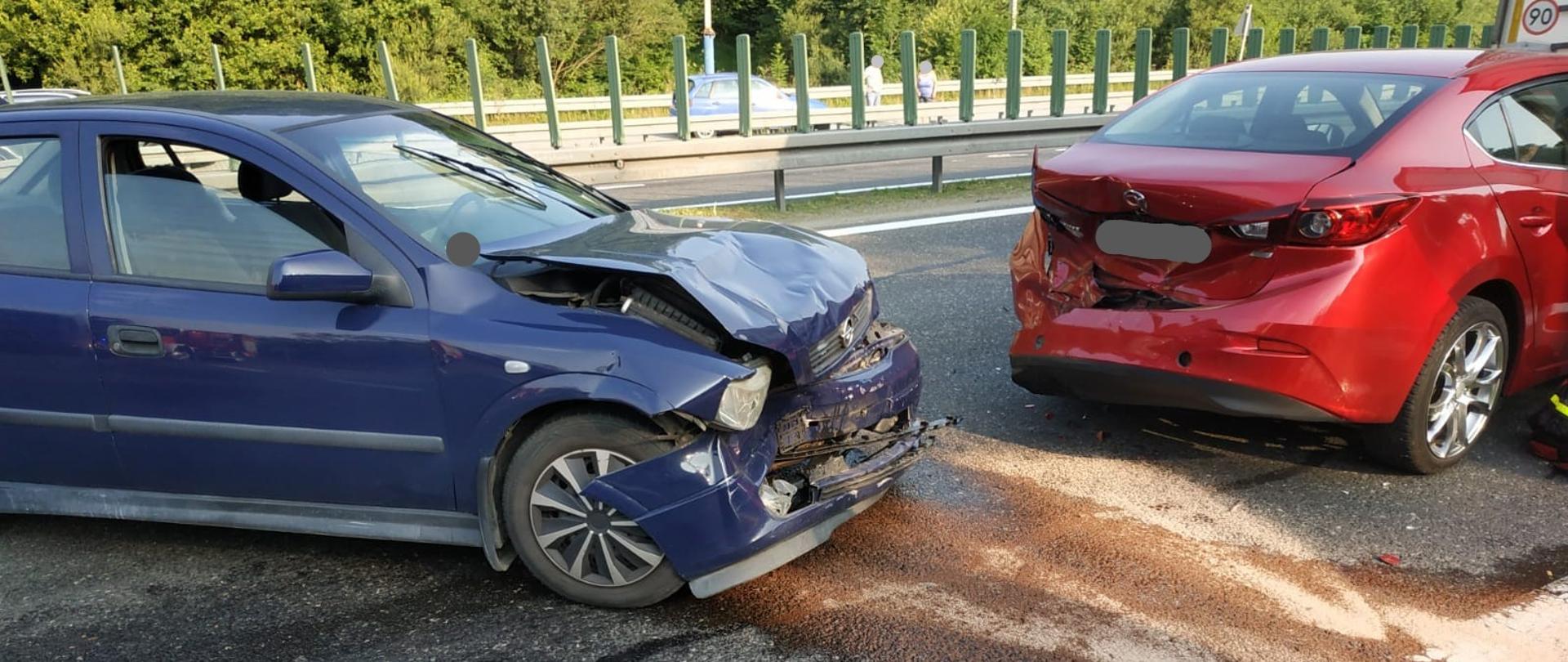 Zdjęcie przedstawia drogę ekspresową na której widzimy dwa samochody osobowe. Jeden w kolorze niebieskim posiada rozbity przód, a drugi w kolorze czerwonym tył. Dodatkowo z prawej strony widoczny jest narożnik naczepy samochodu ciężarowego.