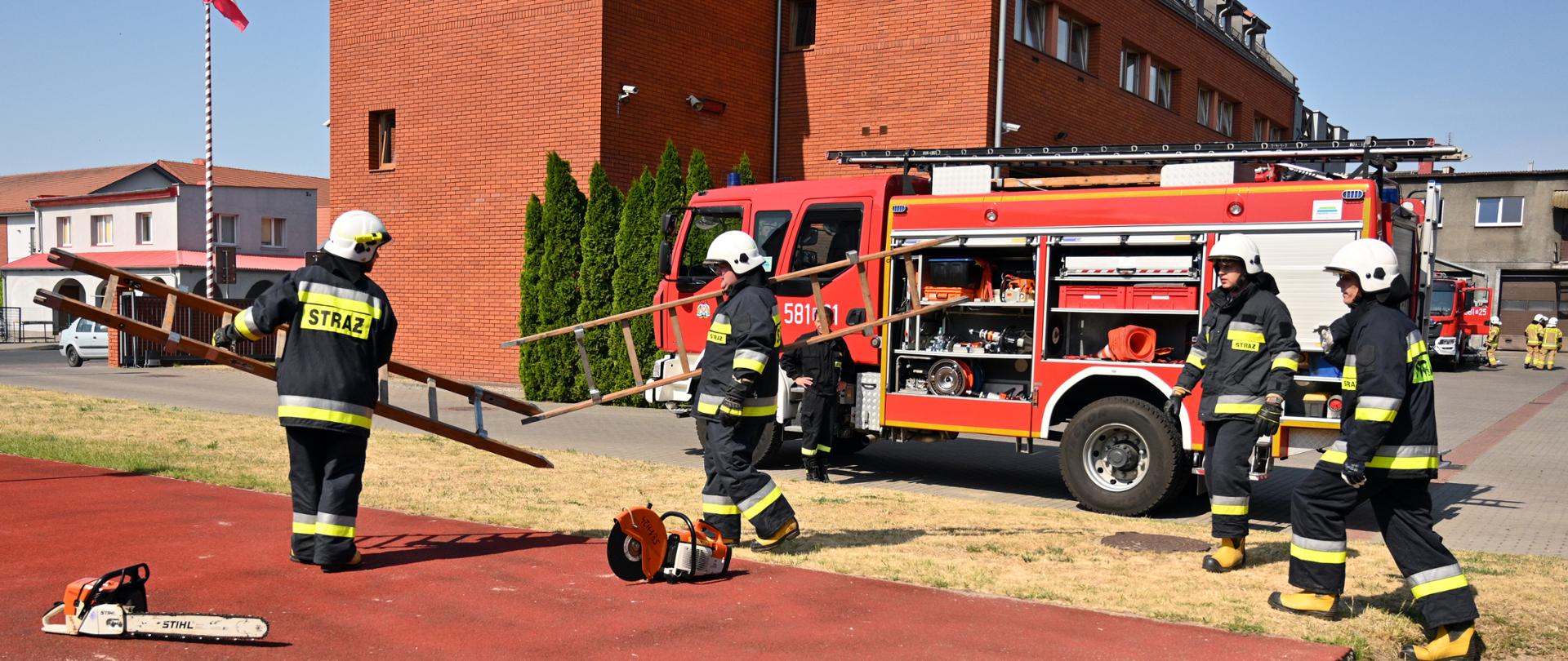 Zdjęcie przedstawia strażaków oraz sprzęt znajdujący się na tartanie dwóch strażaków trzyma po przęśle drabiny za nim samochód pożarniczy z otwartymi skrytkami, za nim budynek z czerwonej cegłu z lewej strony masz z flagą narodową