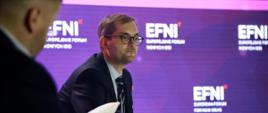Wiceminister rozwoju i technologii Marek Niedużak siedzący podczas obrad EFNI'21 w Sopocie, za jego plecami niebieski baner z logo EFNI