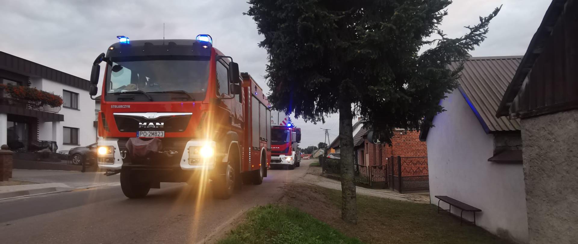 Dwa wozy strażackie z włączonymi światłami ostrzegawczymi ustawione przy budynku podczas akcji gaśniczej, miejsce zabezpieczone pachołkami.