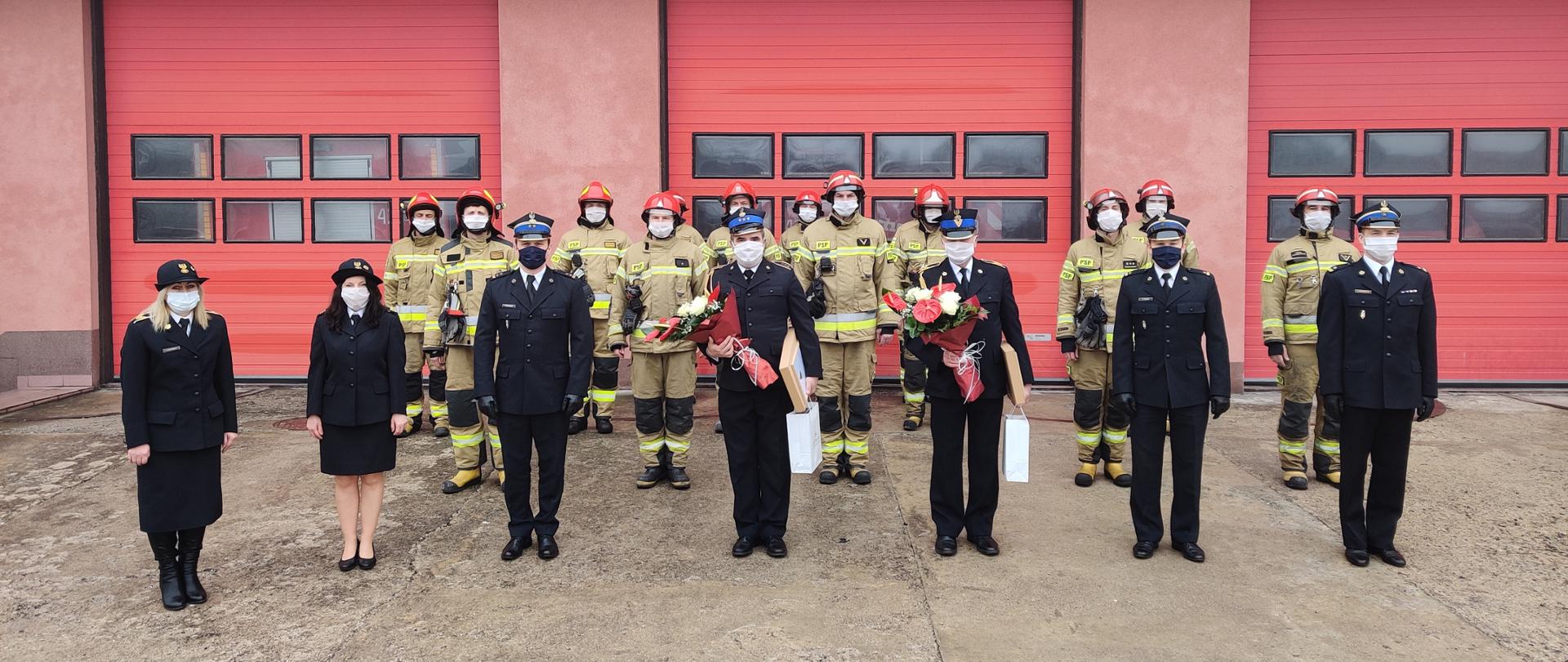 W szyku stoją strażacy w żółtych i granatowych mundurach. W środku dwóch strażaków trzyma kwiaty.
