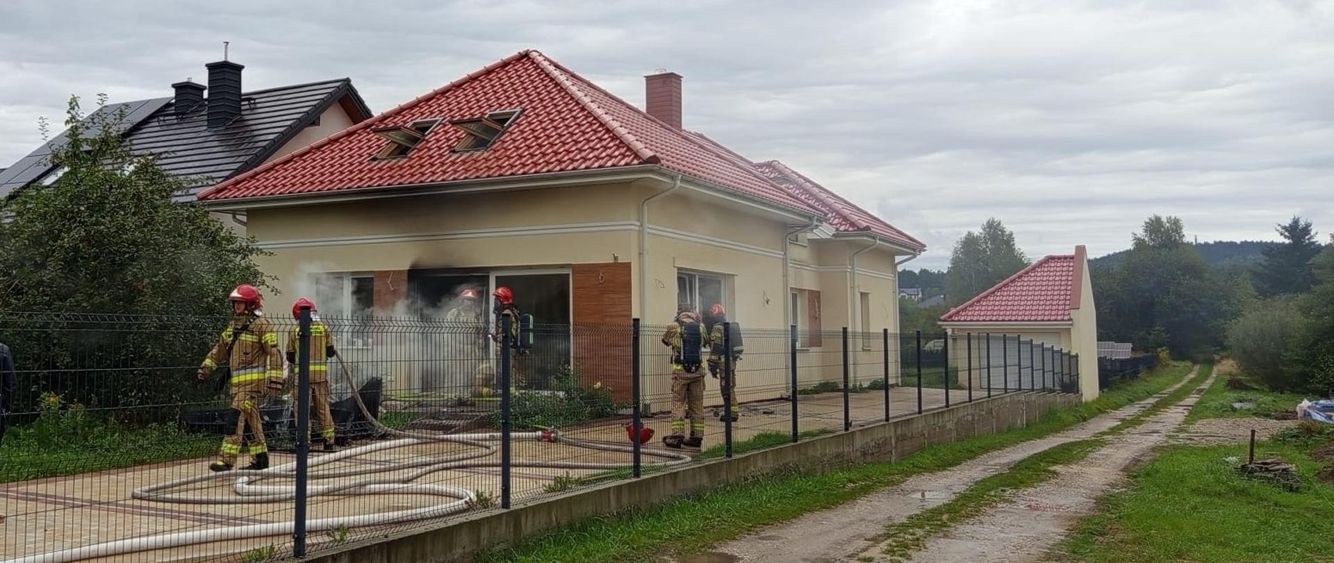 Zdjęcie przedstawia jednorodzinny budynek mieszkalny w którym doszło do pożaru. W otwartych drzwiach tarasowych stoi 2 ratowników zabezpieczonych w aparaty ochrony układu oddechowego, przy budynku kolejnych 4 strażaków kończy działania.