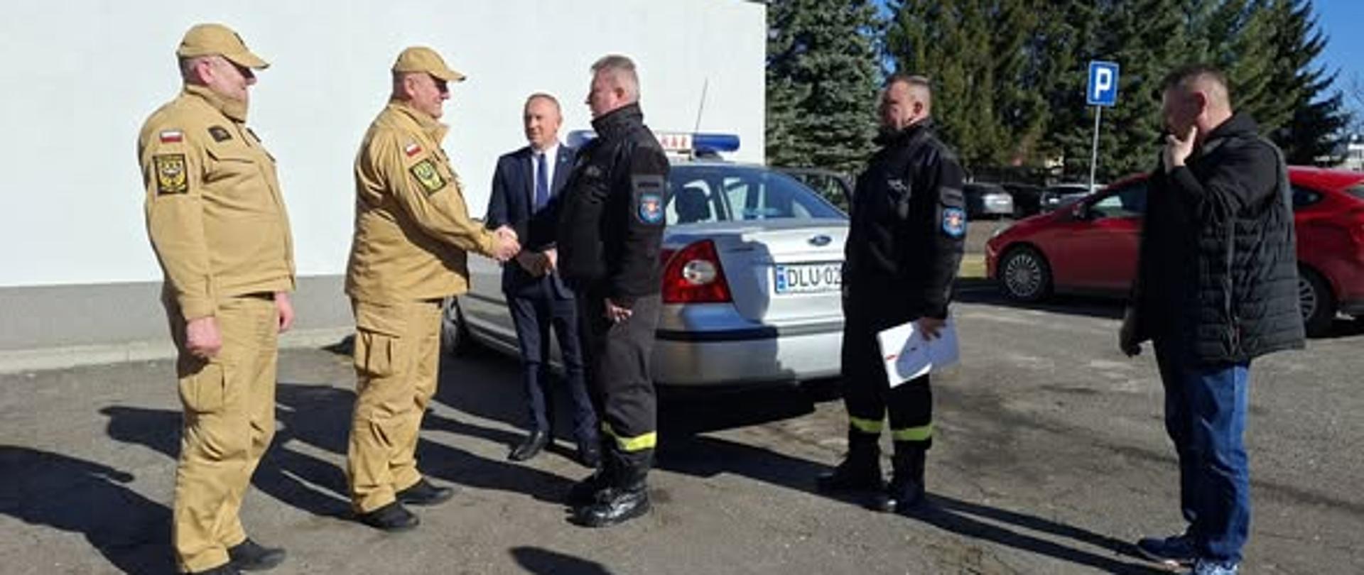 Zdjęcie przedstawia moment przekazania samochodu operacyjnego z Komendy Powiatowej PSP w Lubinie do OSP Chobienia. Na zdjęciu widać sześciu mężczyzn na zewnątrz, na asfaltowym placu, dwóch jest ubranych w mundur służbowy PSP. Spotkanie ma charakter oficjalny – dwóch mężczyzn w centrum uściskiem dłoni przypieczętowuje przekazanie kluczyków do samochodu. W tle widać drzewa iglaste oraz bezchmurne, jasnoniebieskie niebo. Dzień jest słoneczny, na co wskazują wyraźne, długie cienie rzucane przez postacie.