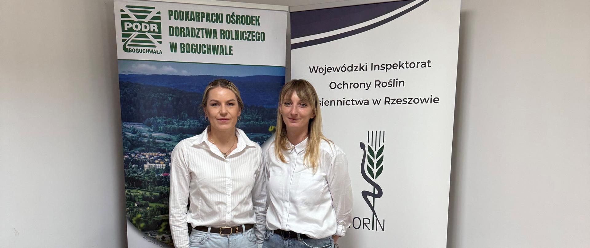 Dwie kobiety w białych koszulach i jasnych dżinsach stoją przy biurku. Na biurku leży laptop i ulotki. Za plecami kobiet stoją banery instytucji.