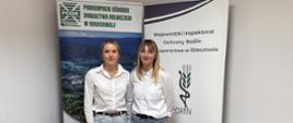 Dwie kobiety w białych koszulach stoją na tle banerów instytucji.