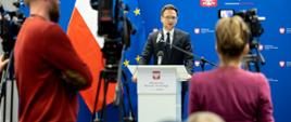 Minister rozwoju i technologii Waldemar Buda na konferencji prasowej, minister stoi za mównicą z mikrofonem, za jego plecami flagi Polski i UE oraz baner z logo MRiT.