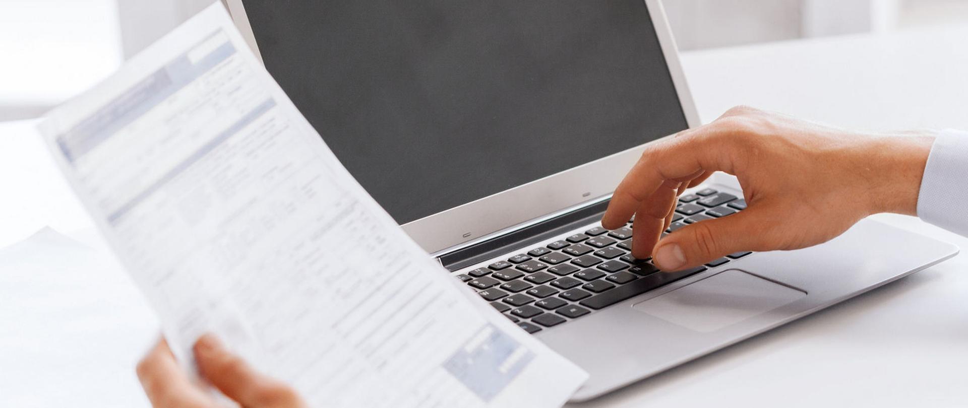 Laptop na biurku oraz jedna dłoń trzyma sprawozdania a druga dotyka klawiatury komputera.