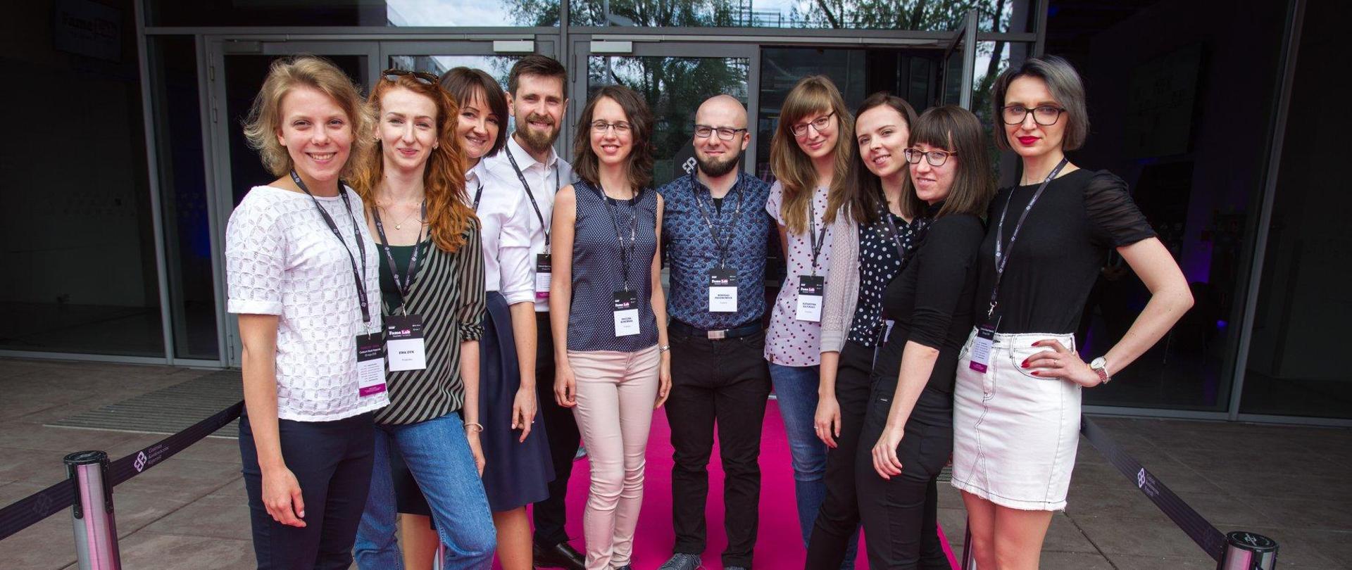 Grupa młodych ludzi stoi na różowym dywanie przed szklanymi drzwiami