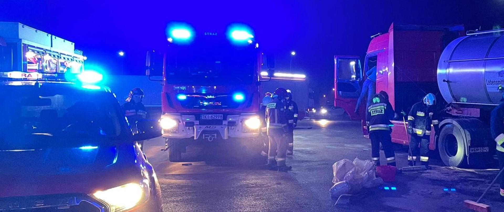 Na zdjęciu z prawej strony znajduje się samochód ciężarowy w którym doszło do wycieku oleju napędowego. Widać tam strażaków usuwających wyciek za pomocą sorbentu. Z lewej strony znajdują się samochody pożarnicze – całe zdjęcie ma niebieską poświatę.