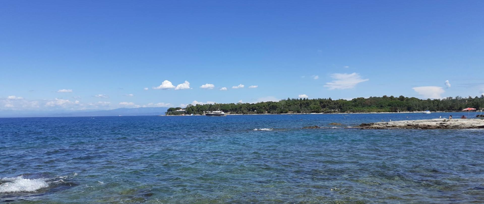 Fotografia przedstawia widok na morze, w piękny bezchmurny dzień, w oddali fragment zalesionej wyspy.