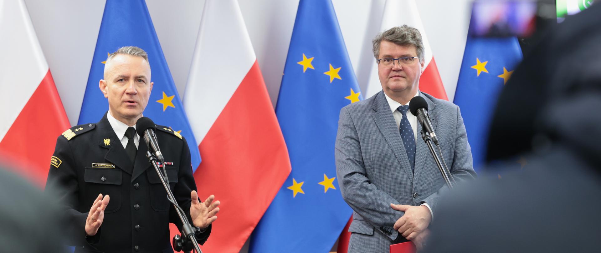 Na zdjęciu Komendant Główny PSP w czarnym mundurze wyjściowym po prawej Minister Wąsik z szarym mundurze. W tle Flagi biało czerwone oraz flaga Unii Europejskiej niebieska z żółtymi gwiazdkami.