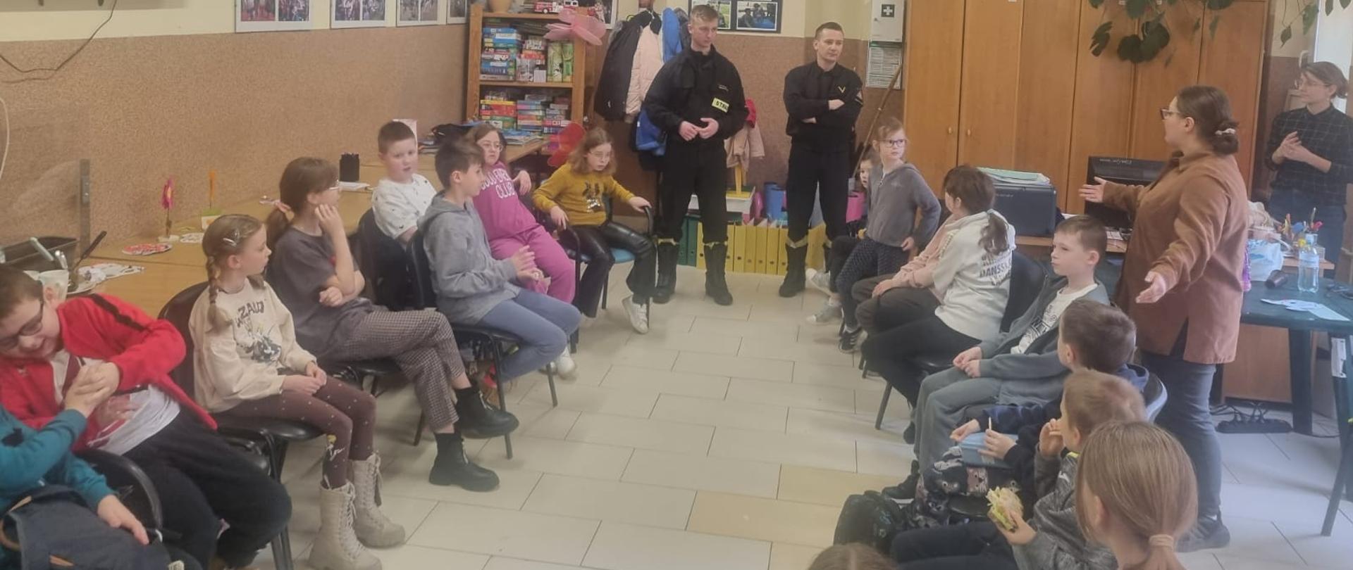 Na prawo i na lewo na 1 planie siedzą dzieci w rzędzie, na prosto w tle stoi dwóch strażaków, w tle na prawo szafy oraz opiekunka. Na podłodze płytki . na lewo na ścianie wisi telewizor a za nim zdjęcia. 