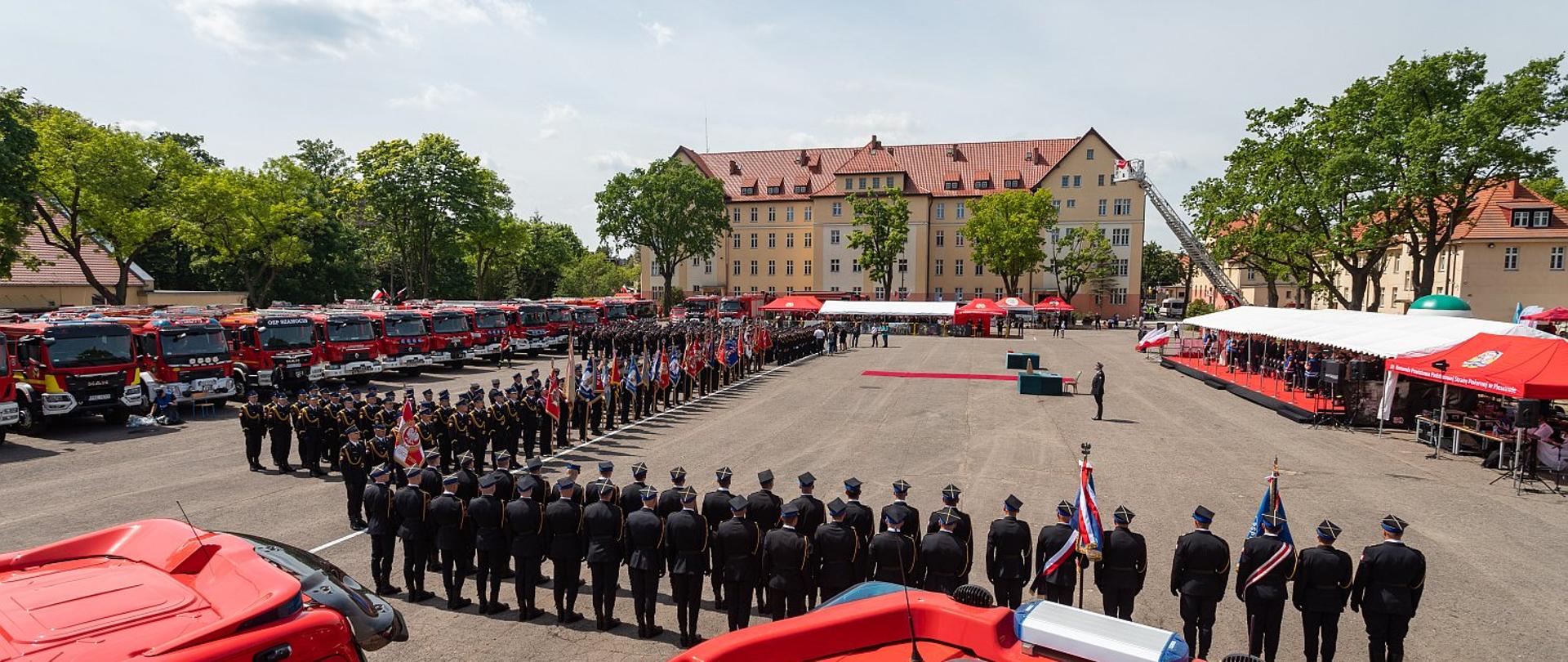 Na zdjęciu widać plac na którym znajdują się funkcjonariusze w mundurach wyjściowych. Zdjęcie wykonane z samochodu ratowniczo-gaśniczego. Kolor czerwony. Zdjęcie zrobione w ciągu dnia. Niebo błękitne. W tle budynek ze spadzistym dachem z dachówki. Po lewej stronie zielone drzewa.