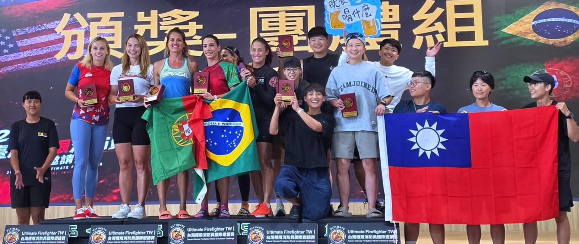 Grupa zawodniczek stojących na podium oraz obok niego podczas ceremonii wręczenia nagród zawodów „Ultimate Firefighter TWI”. Uczestniczki trzymają statuetki i flagi Portugalii, Brazylii oraz Tajwanu.