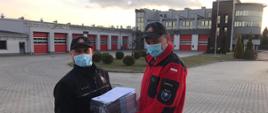 Przekazanie ulotek informacyjnych na temat profilaktyki oraz szczepień przeciwko COVID-19 dla druhów ochotniczych straży pożarnych powiatu lublinieckiego. Strażak państwowej straży pożarnej przekazuje pakiet ulotek dla druha ochotniczej straży pożarnej. W tle budynek komendy PSP.