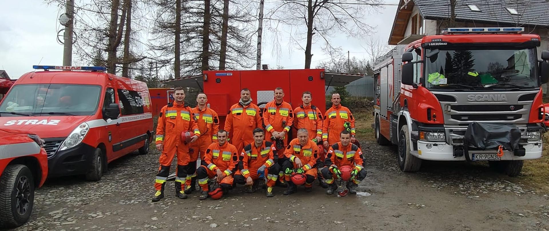 Strażacy ubrani w pomarańczowe kombinezony bez nakrycia głowy, ustawienie w dwóch rzędach, obok samochody pożarnicze z tyłu drzewa