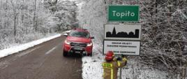 Na drodze obok zaśnieżonego lasu strażak obok czerwonego wozu bojowego przykręcają tablicę informacyjną.