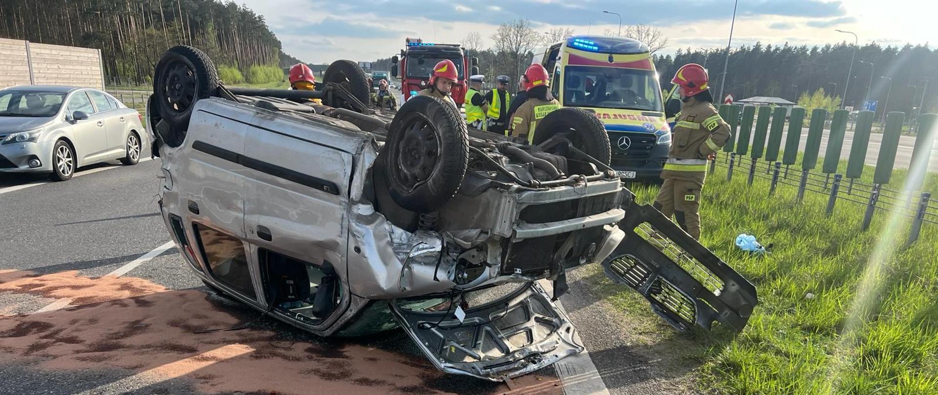 Zdjęcie przedstawia rozbity samochód osobowy znajdujący się na dachu. Na zdjęciu widać strażaków w umundurowaniu specjalnym oraz funkcjonariuszy policji. W tle widać karetkę pogotowia oraz pojazd pożarniczy.