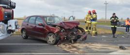 Kolizja na DW434. Skrzyżowanie dróg w okolicach Roszkówka. Na jezdni stoi rozbity ford. Auto ma mocno zniszczony przód. Z prawej strony znajduje się 3 strażaków. Z lewej strony widoczny jest fragment wozu strażackiego. W tle pola, drzewa i zabudowania.