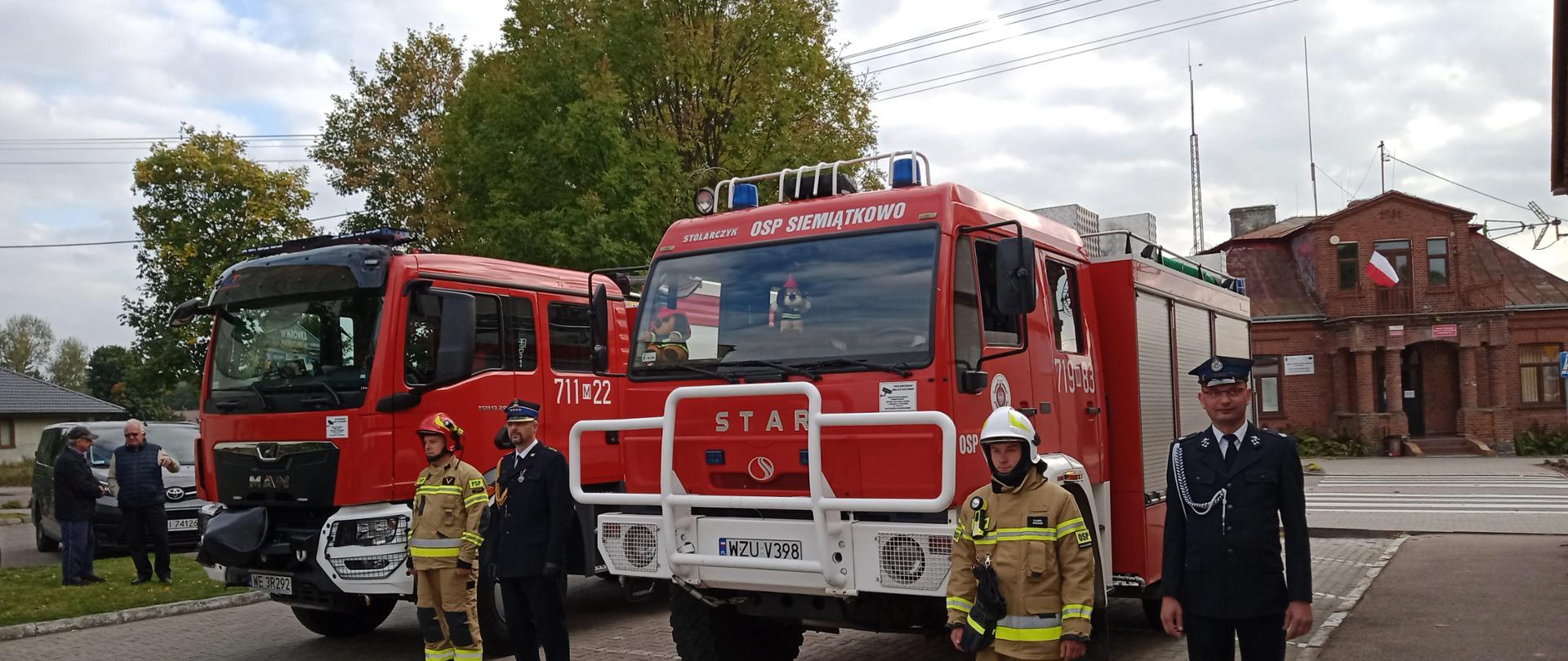 Dwa samochody ratowniczo- gaśnicze stojące na placu przed budynkiem Gminy. Obok samochodów stoją straży PSP oraz OSP. W tle budynki i drzewa.