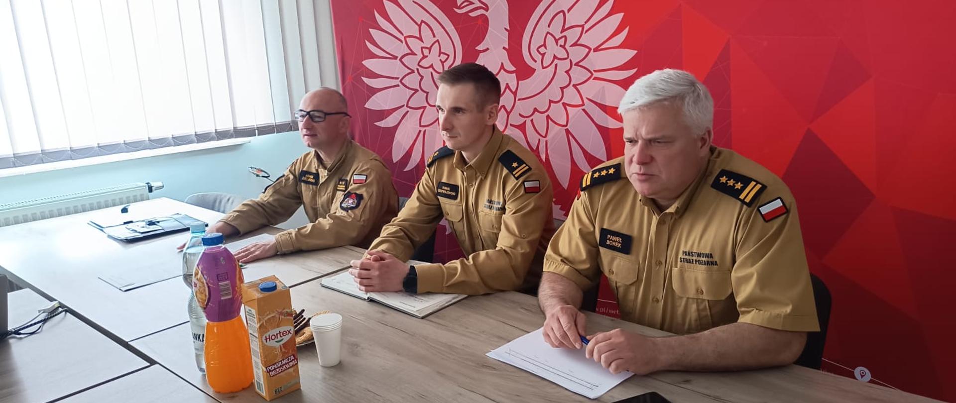 Przy stole konferencyjny na tle czerwonej ścianki z białym godłem siedzą trzej funkcjonariusze Państwowej Straży Pożarnej