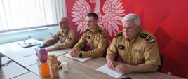 Przy stole konferencyjny na tle czerwonej ścianki z białym godłem siedzą trzej funkcjonariusze Państwowej Straży Pożarnej