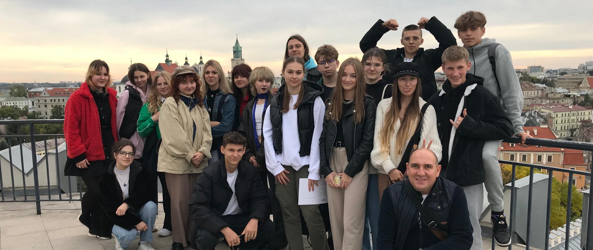 Na zdjęciu kilkunastoosobowa grupa młodzieży wraz z opiekunem znajduje się na tarasie widokowym, w tle panorama Lublina