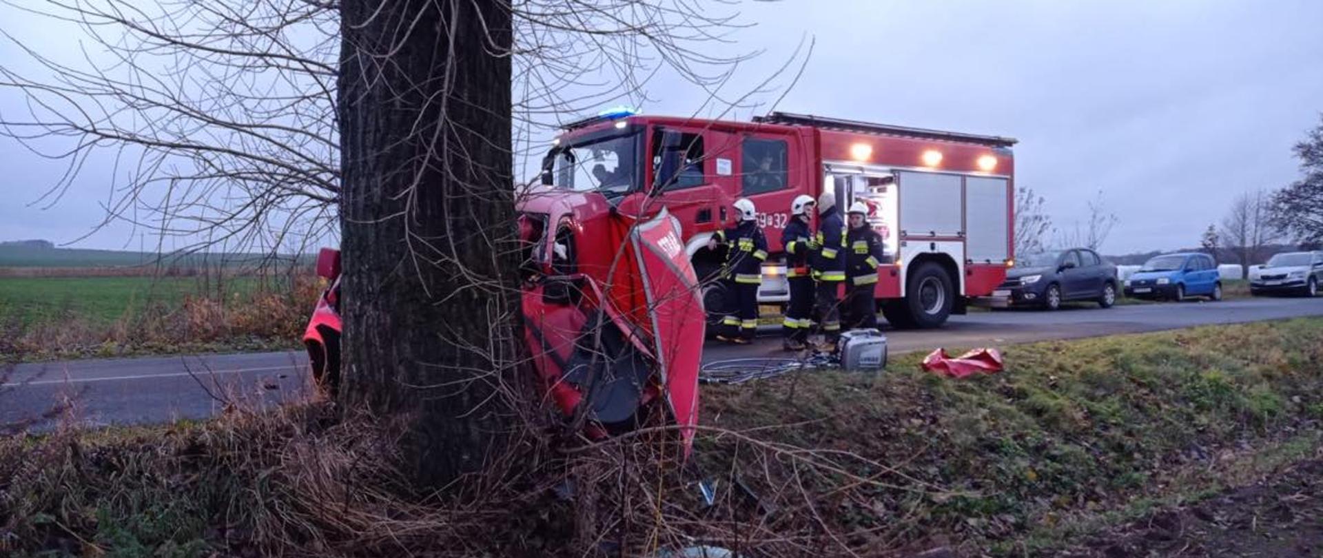 Zdjęcie przedstawia samochód osobowy, który uderzył w drzewo. W tle widać strażaków. Jest dzień, pochmurno, bez opadów. 