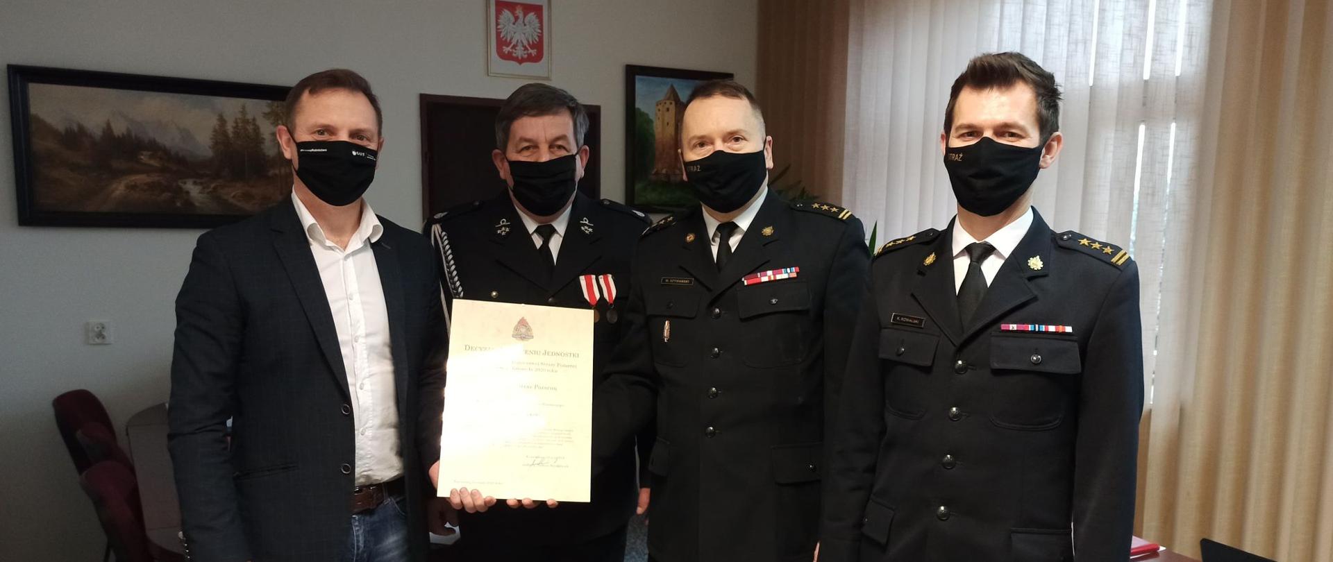 Zdjęcie przedstawia czterech mężczyzn, stojących obok siebie, zwróconych przodem do obiektywu. Mężczyźni znajdują się w gabinecie wójta. Pierwszy od lewej mężczyzna (wójt) ubrany jest w dżinsy, białą koszulę i grafitową marynarkę. Kolejny to Prezes OSP Regnów ubrany jest w mundur galowy i w dłoniach trzyma ramkę z decyzja o włączenie do KSRG. Kolejni dwaj mężczyźni to komendant i z-ca komendanta KP PSP w Rawie Maz. ubrani w galowy mundur. Wszyscy Panowie mają na sobie czarne maseczki.
