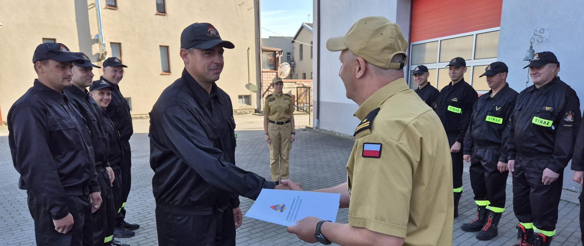 Plac komendy powiatowej państwowej straży pożarnej w sępólnie krajeńskim, strażacy dwóch zmian służbowych w ubraniach koszarowych stoją w szeregu naprzeciwko siebie, między nimi zastępca komendanta powiatowego młodszy brygadier Robert Liss gratuluje wyróżnionemu strażakowi.