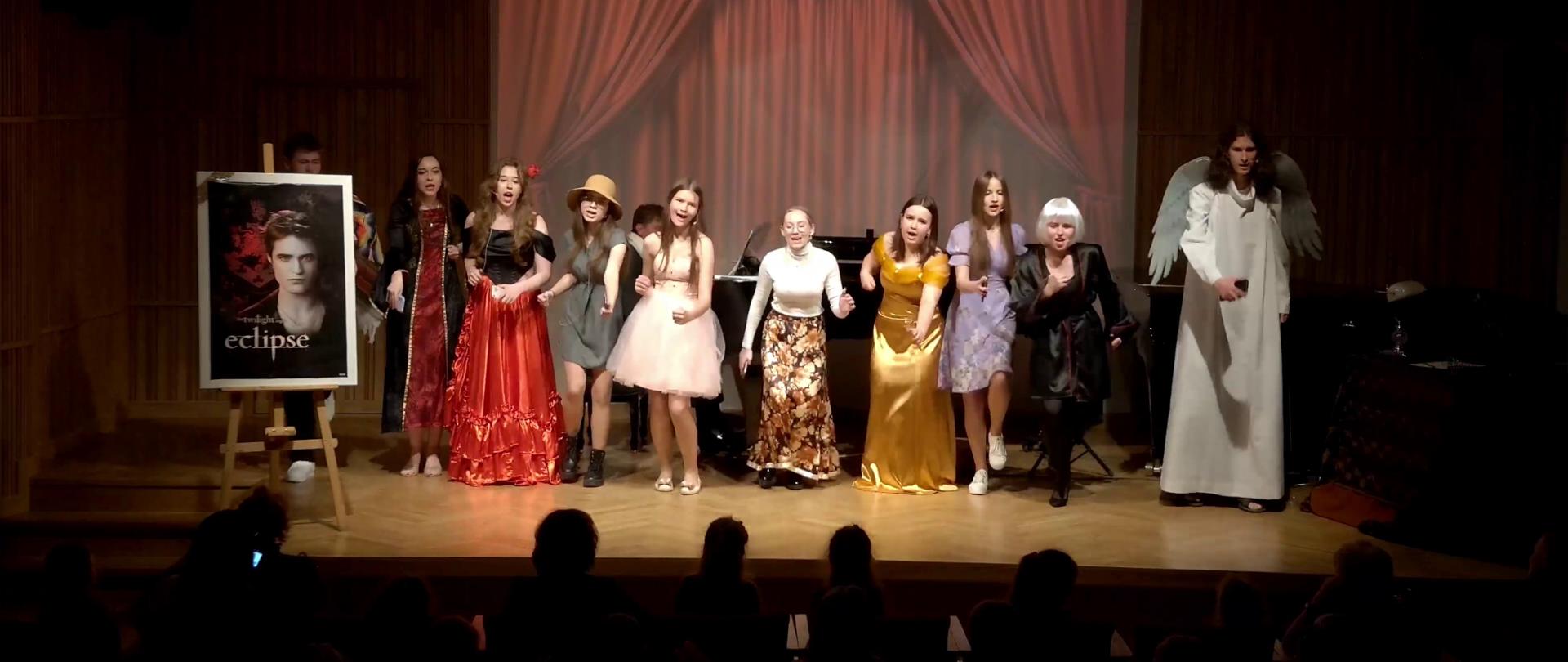 Zdjęcie. estrada sali koncertowej. Śpiewają i tańczą uczniowie. Przed nimi siedzą widzowie. W tle wyświetlona jest grafika przedstawiająca kurtynę teatru.