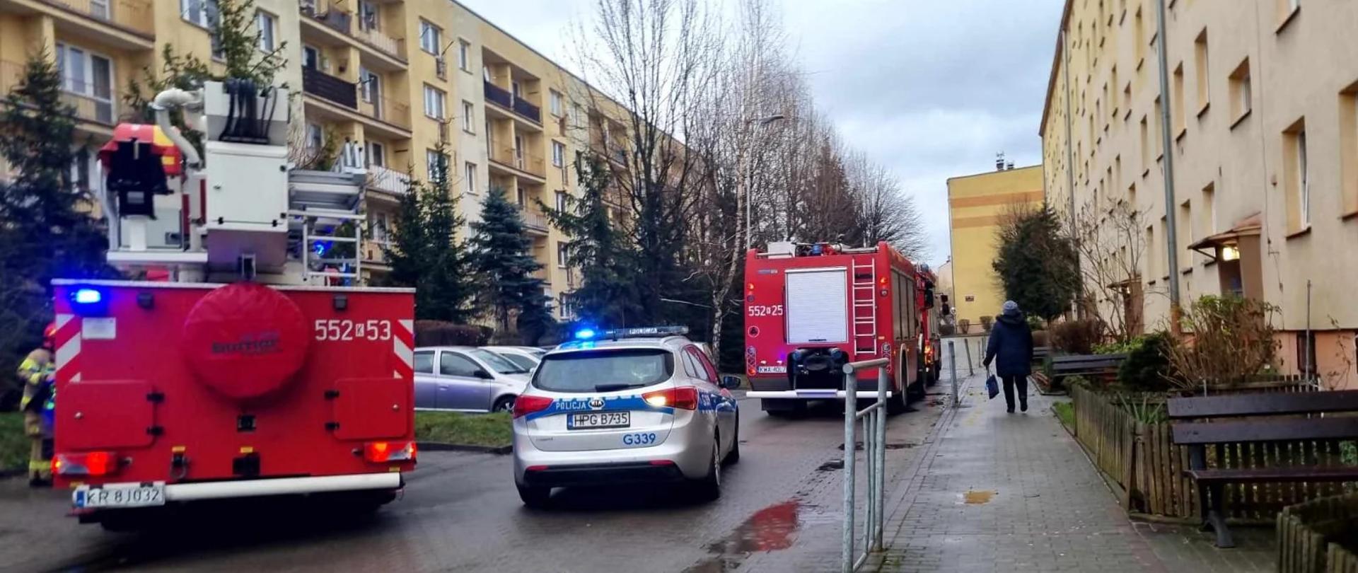 Droga między blokami. Na niej trzy samochody strażackie i radiowóz. Skrajnie po lewej widoczny strażak. Po prawej widoczna od tyłu osoba idąca wzdłuż bloku.
