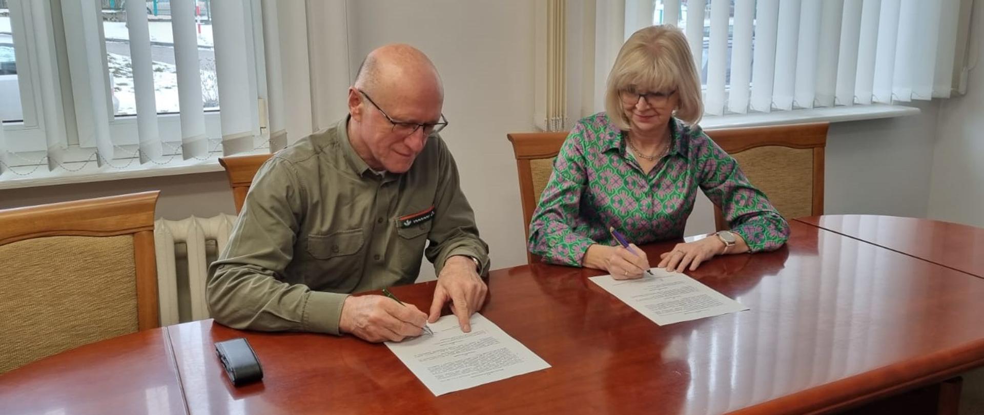 Przy stole siedzi Regionalny Dyrektor Ochrony Środowiska w Olsztynie Agata Moździerz i Dyrektor Regionalnej Dyrekcji Lasów Państwowych w Białymstoku Tadeusz Wilczyński. Na stole leżą dwa dokumenty. W rękach trzymają długopisy.