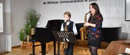 Na zdjęciu wykonanym w auli PSM, widnieje uczeń grający na saksofonie wraz z nauczycielem saksofonu Agatą Bartoš. Na pierwszym planie widać fragment organów, w tle fortepian. Kolorystyka zdjęcia jest biało-czarno-brązowa.