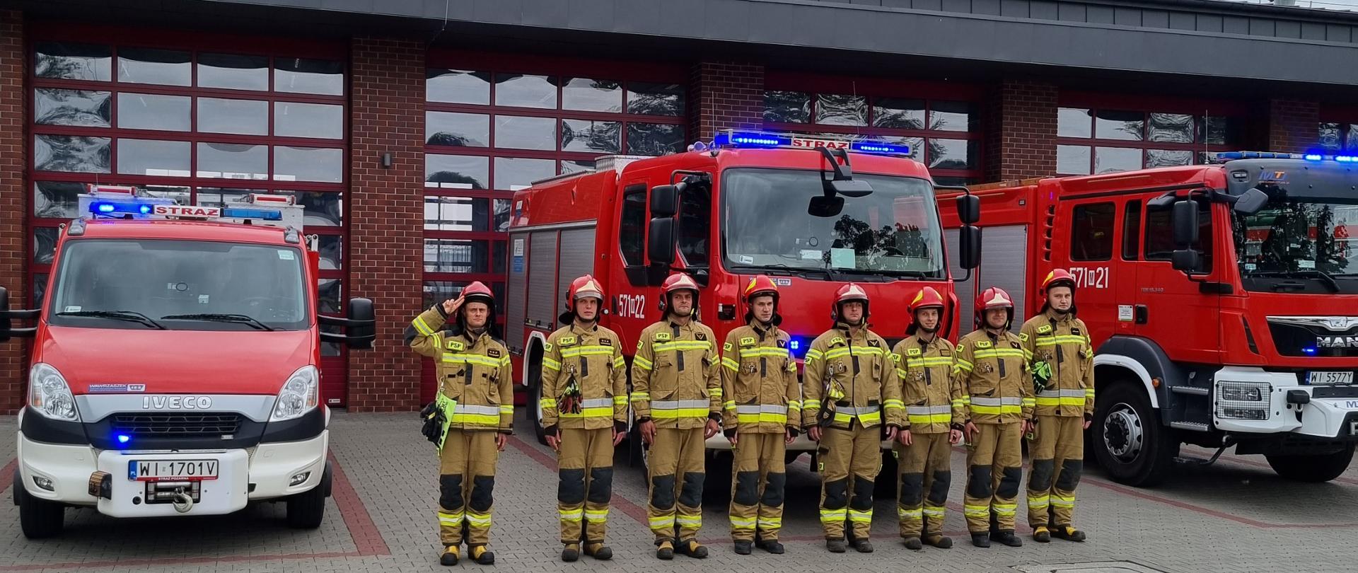Pododdział strażaków ustawiony w dwuszeregu, w ubraniach specjalnych koloru piaskowego. Dowódca salutuje. W tle samochody strazackie oraz fragment budynku.
