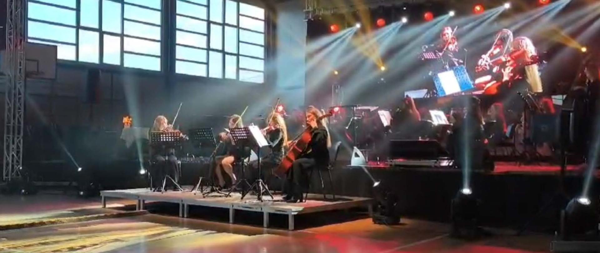 Muzycy Orkiestry Kameralnej PSM II stopnia w Mielcu wraz z dyrygentem stoją na scenie podczas koncertu „Muzyka ma twarz kobiety” w sali widowiskowej w Czerminie.