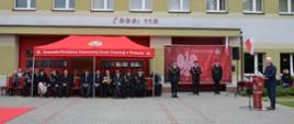 uczestnicy uroczystości siedzą pod namiotem wystawowym obok stoją strażacy, za nimi budynek komendy powiatowej