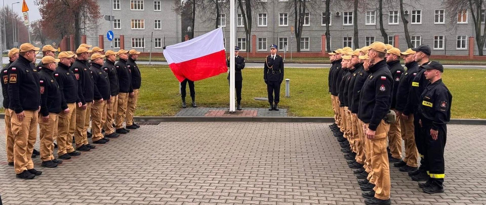 Na placu przed Komendą Miejska trwa podnoszenie białoczerwonej flagi na maszt. Po obu stronach masztu, w mundurach, stoją na przeciwko siebie funkcjonariusze w mundurach służbowych. w tle zabudowania miejskie.