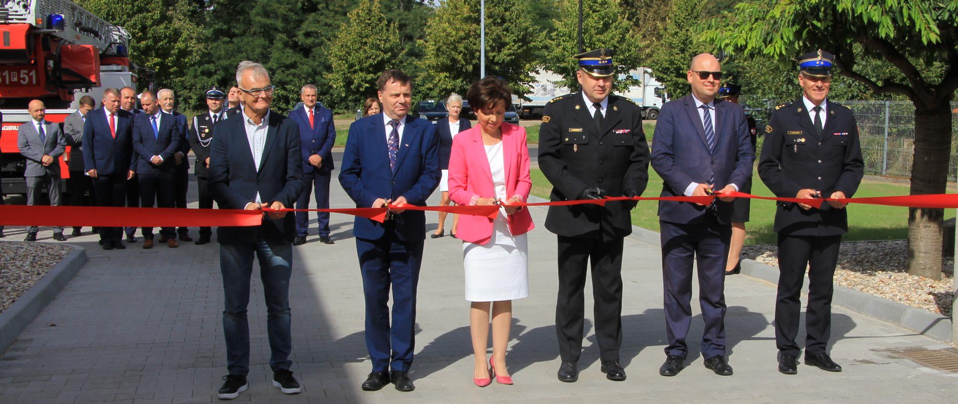 grupa ludzi na dużym placu przecina czerwoną wstęgę, otwarcie, w tle drzewa oraz duża grupa ludzi i samochód pożarniczy