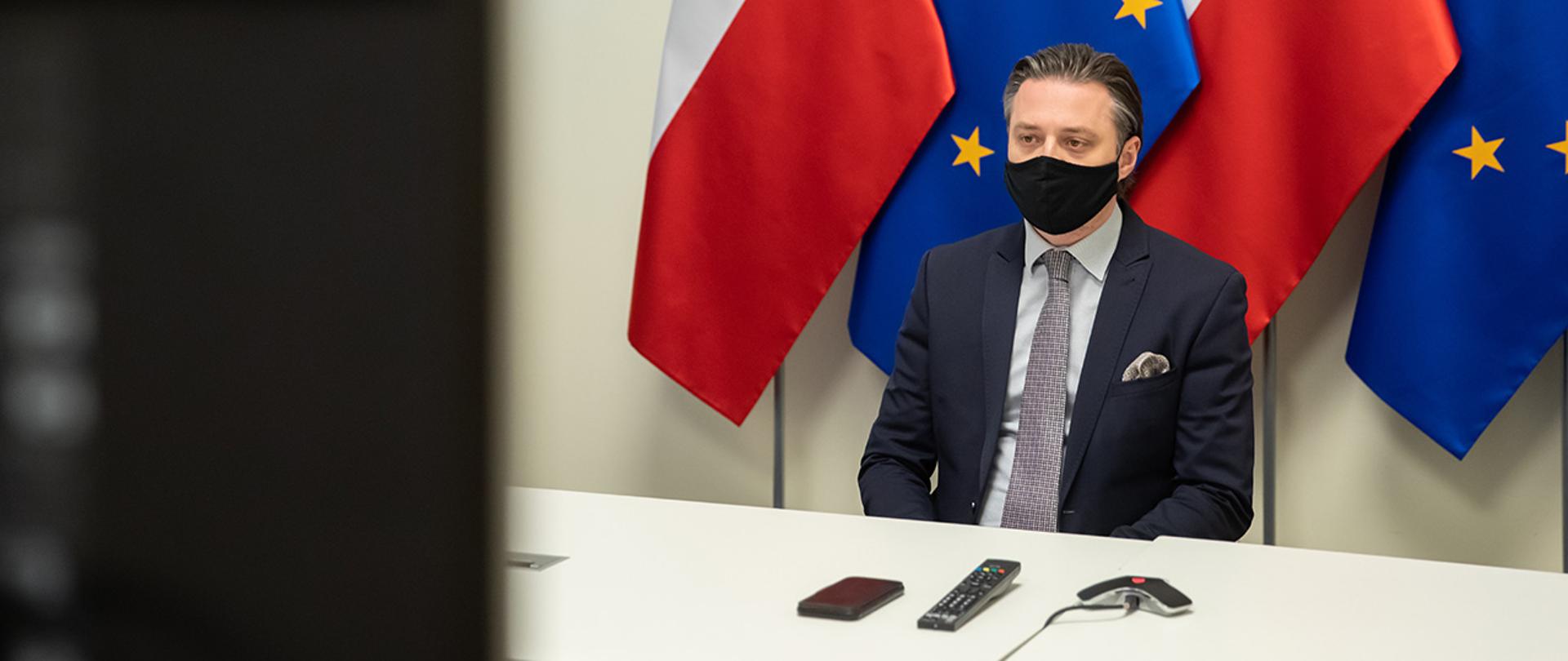 Na zdjęciu widać wiceministra Bartosza Grodeckiego siedzącego za stołem na tle flag Polski i UE wpatrzonego w ekran.