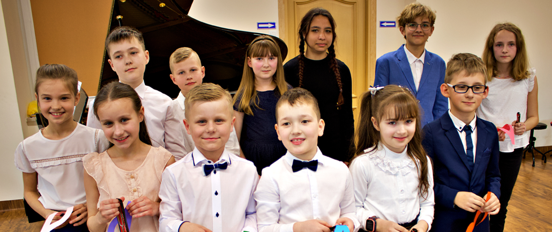 Na zdjęciu grupa 12 dzieci, dziewczynki i chłopcy,. stoją na tle fortepianu, w rękach trzymają kolorowe kotyliony i małe kamertony.